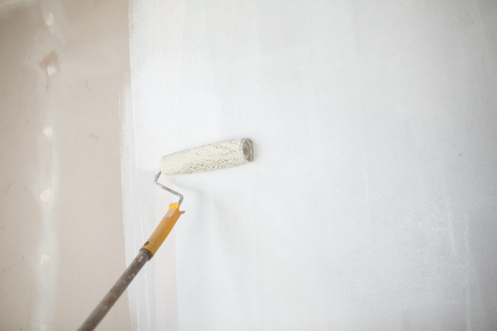 A person uses a brush roller to paint drywall white